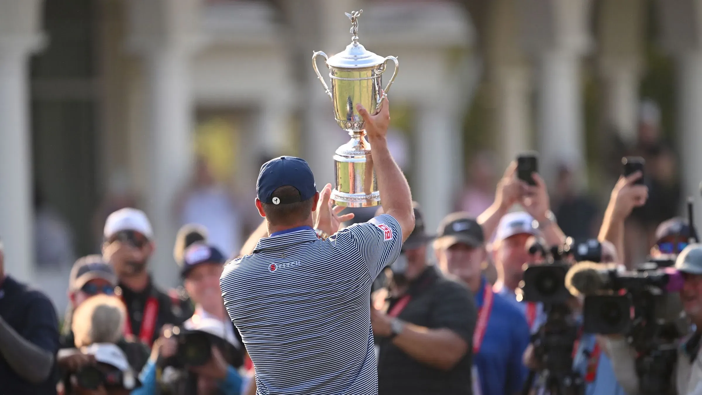 لاعب غولف يحمل الكأس في احتفال بعد الفوز، محاط بالجماهير والكاميرات، في بطولة الولايات المتحدة المفتوحة.