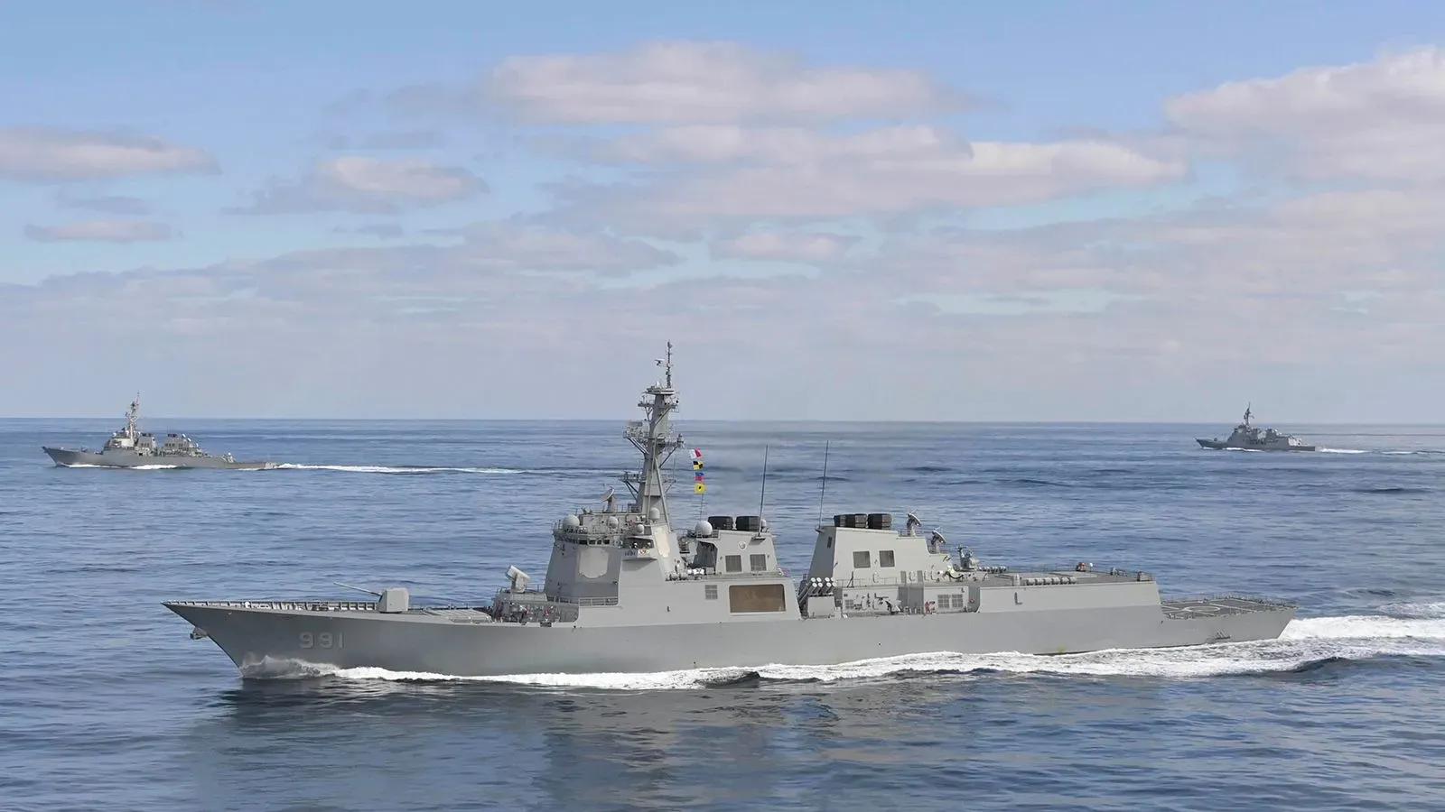 سفينة حربية حديثة تبحر في مياه مفتوحة، محاطة بسفينتين أخريين، تعكس التعاون بين الولايات المتحدة وكوريا الجنوبية في بناء السفن.