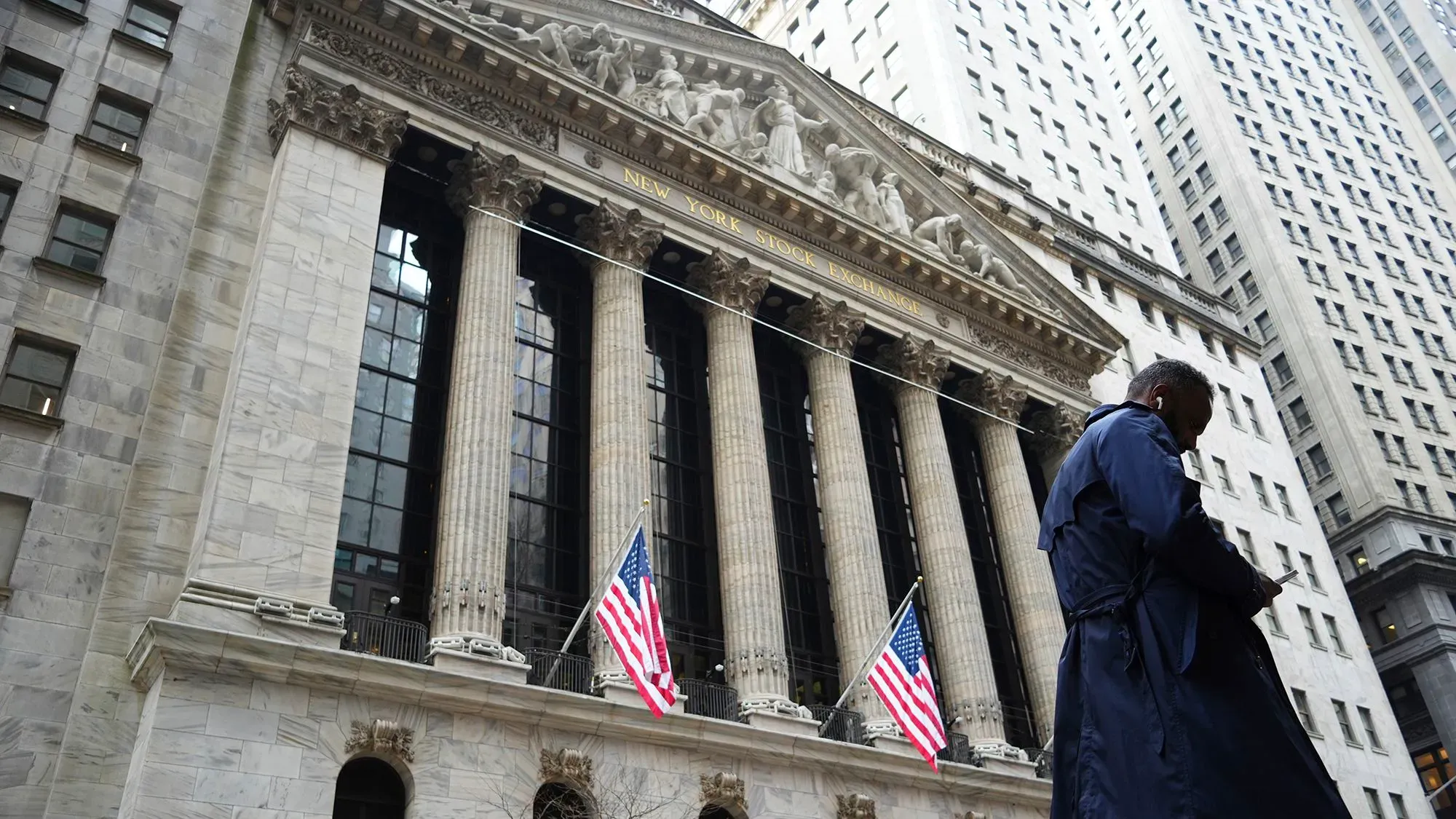 واجهة بورصة نيويورك مع أعلام أمريكية ورجل يرتدي معطفًا، تعكس حالة السوق المتقلبة والهبوط الحاد في الأسهم.