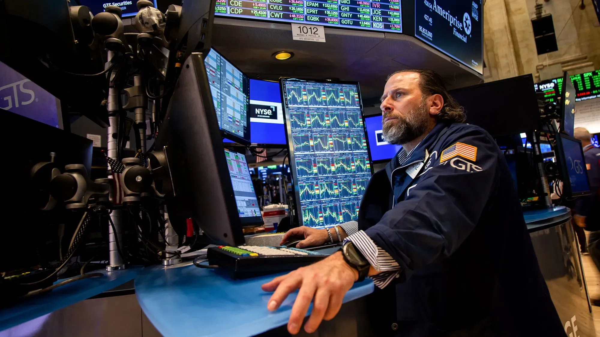 تاجر في سوق الأسهم الأمريكية يتابع شاشات متعددة تعرض بيانات السوق، مع وجود علامات على التقلبات الاقتصادية.