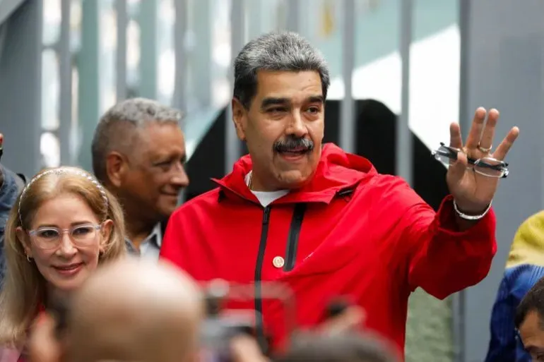 مادورو، الرئيس الفنزويلي، يلوح بيده خلال حدث عام، مرتديًا سترة حمراء، مع وجود أشخاص آخرين في الخلفية.