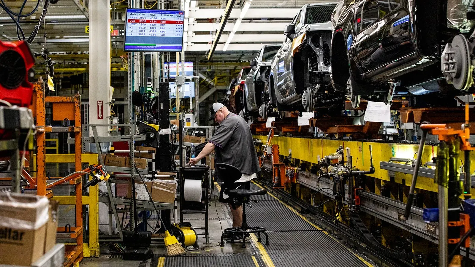 عامل يعمل في مصنع لتجميع السيارات، مع وجود شاشات عرض توضح البيانات الإنتاجية، مما يعكس تأثير التضخم على الصناعة.