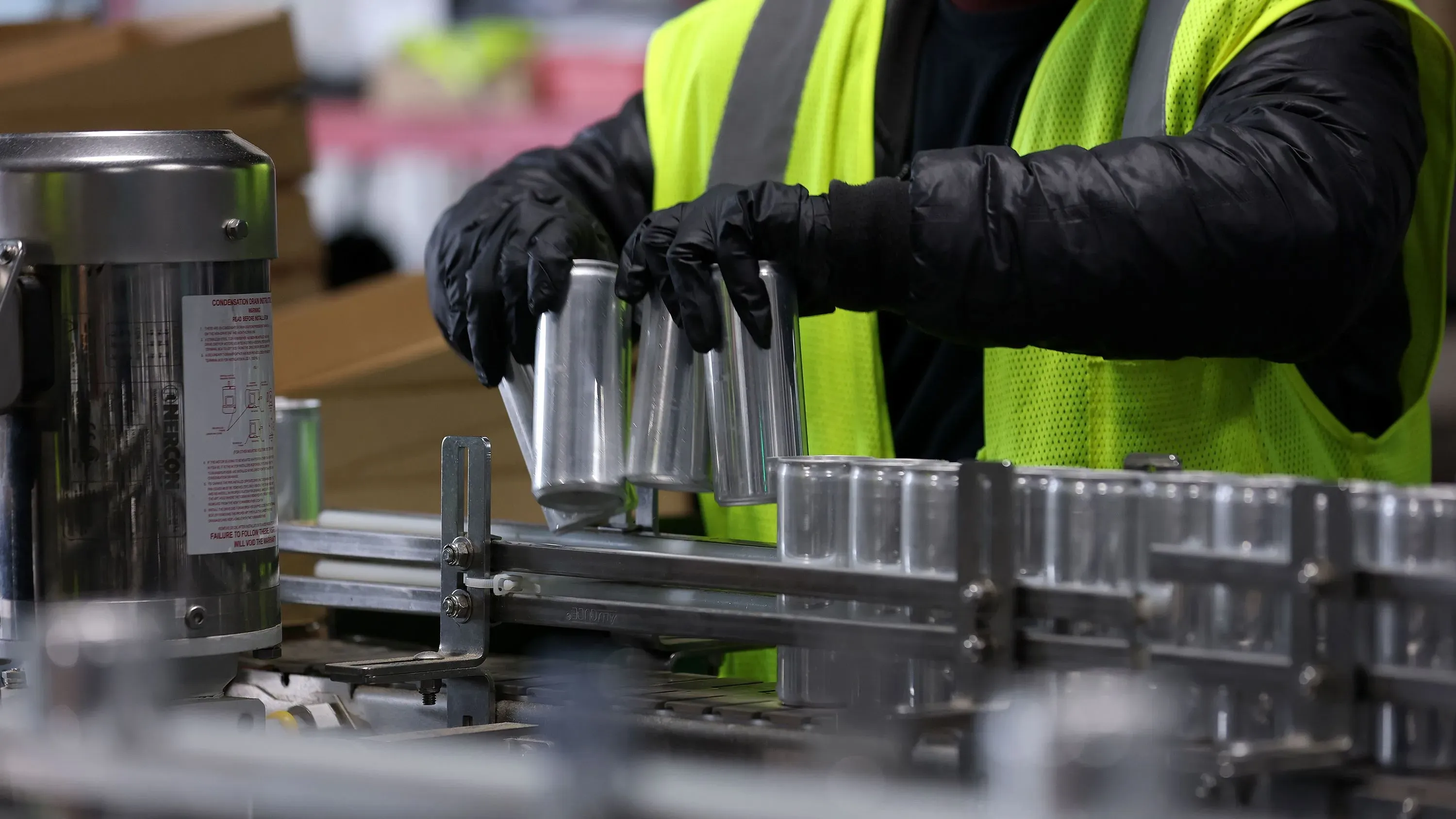 عامل يرتدي سترة عاكسة وقفازات، يقوم بتجميع علب معدنية على خط إنتاج، في سياق انخفاض أسعار الجملة في الولايات المتحدة.