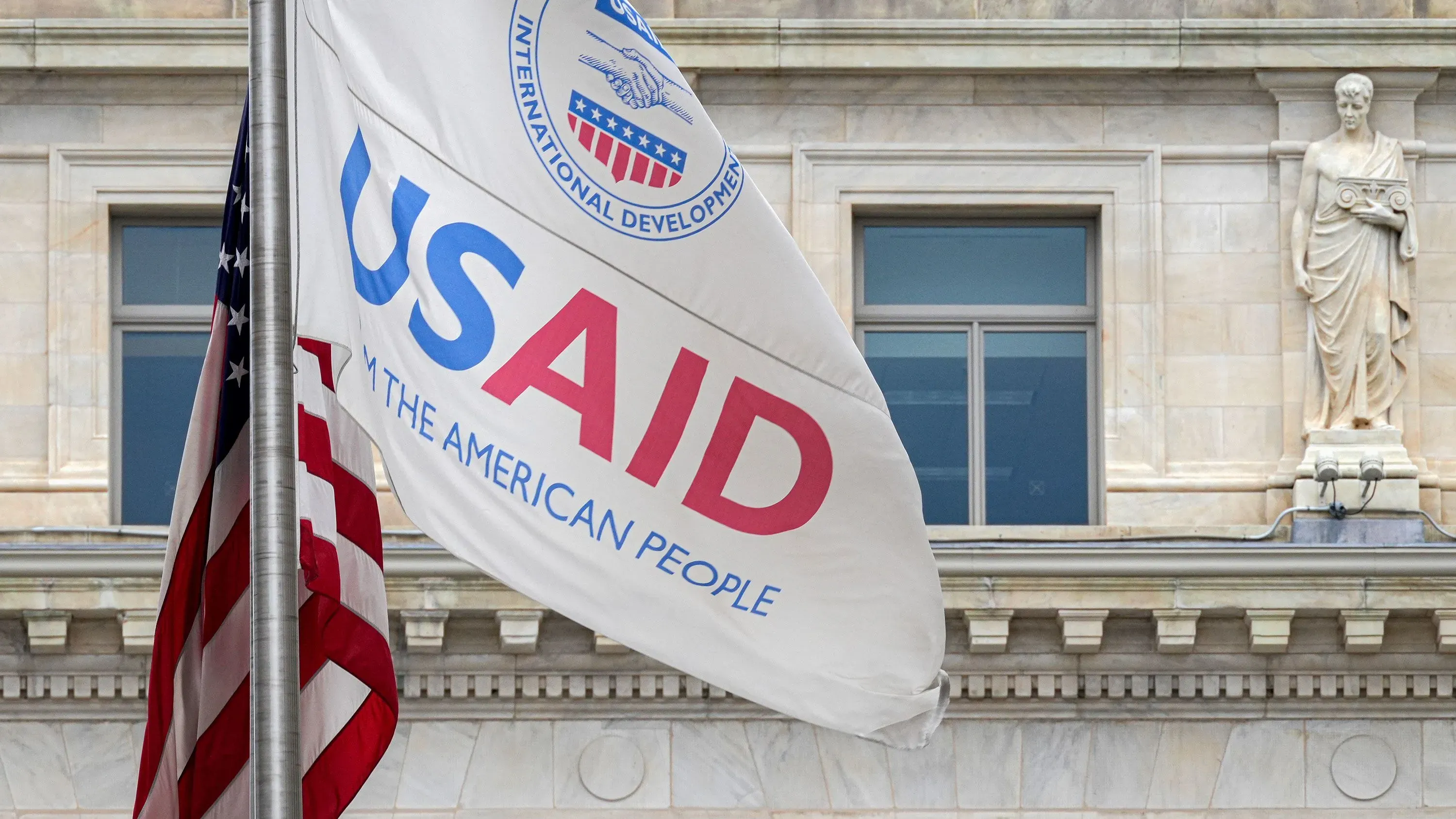 علم الوكالة الأمريكية للتنمية الدولية (USAID) يتدلى بجانب العلم الأمريكي، مع واجهة مبنى تاريخي في الخلفية، مما يعكس تأثير الوكالة في تقديم المساعدات الإنسانية.