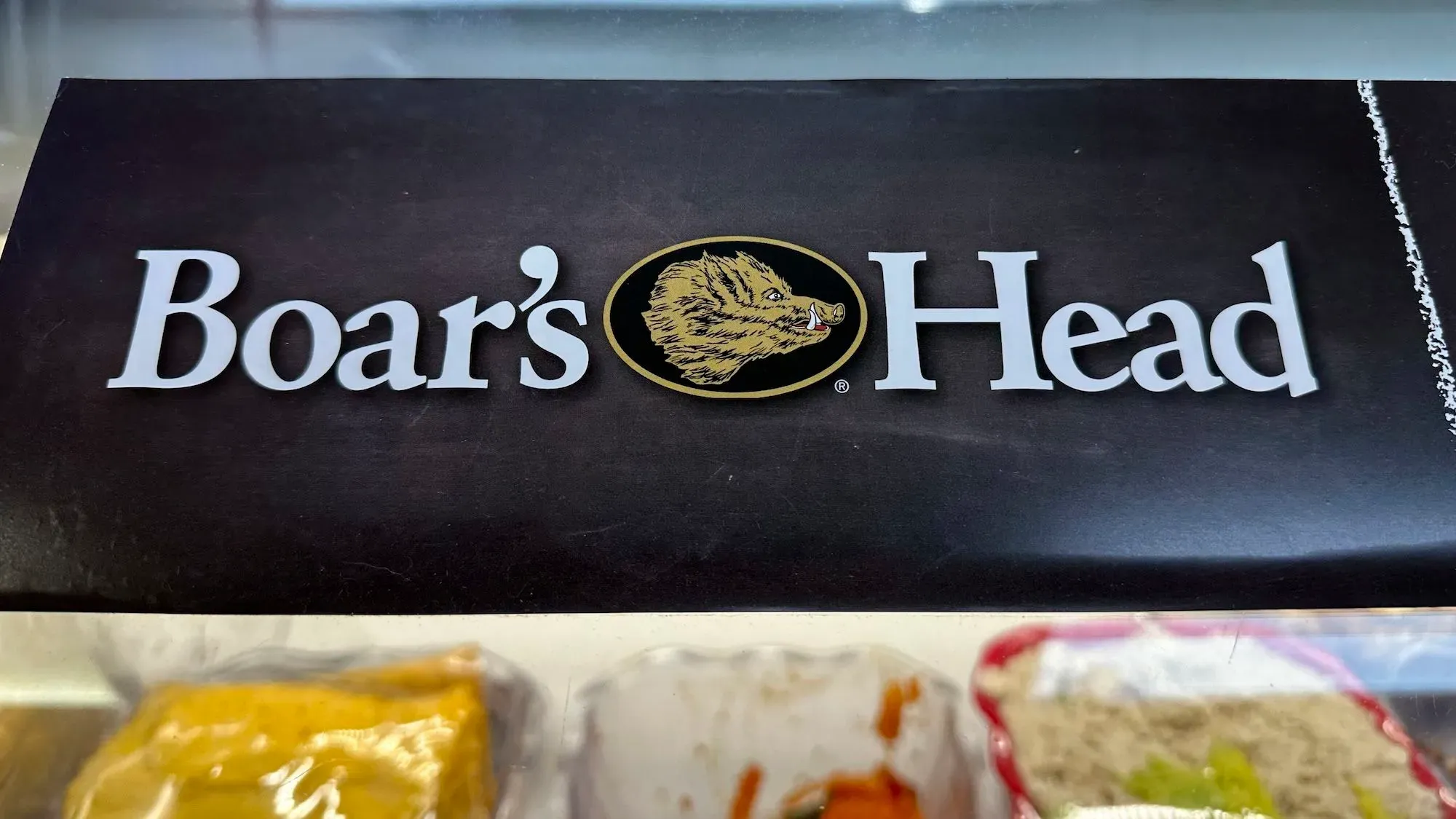 شعار شركة Boar's Head يظهر في الصورة، مع خلفية داكنة وعبوات متنوعة من اللحوم الجاهزة، مما يعكس القضايا الصحية المثارة حول مصانع الشركة.