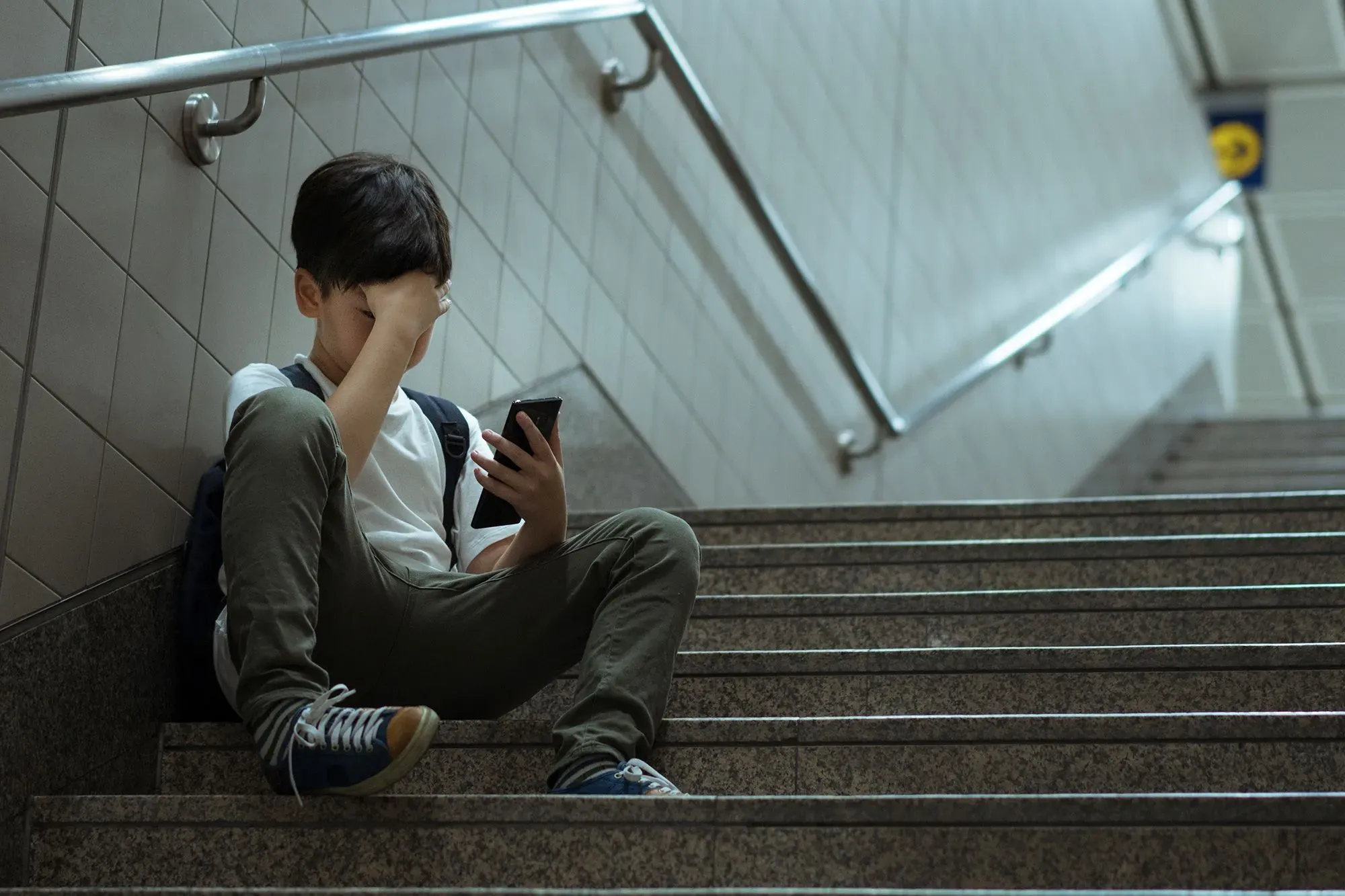 طفل يجلس على الدرج، ممسكًا بهاتفه، ويبدو عليه الإحباط أو القلق، مما يعكس تأثير وسائل التواصل الاجتماعي على الصحة النفسية.