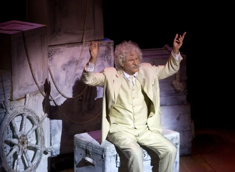 مسرحية تُظهر شخصية ترتدي ملابس بيضاء، تجلس على صندوق وتقوم بإيماءات تعبيرية، تعكس روح الفكاهة والأداء الفني.