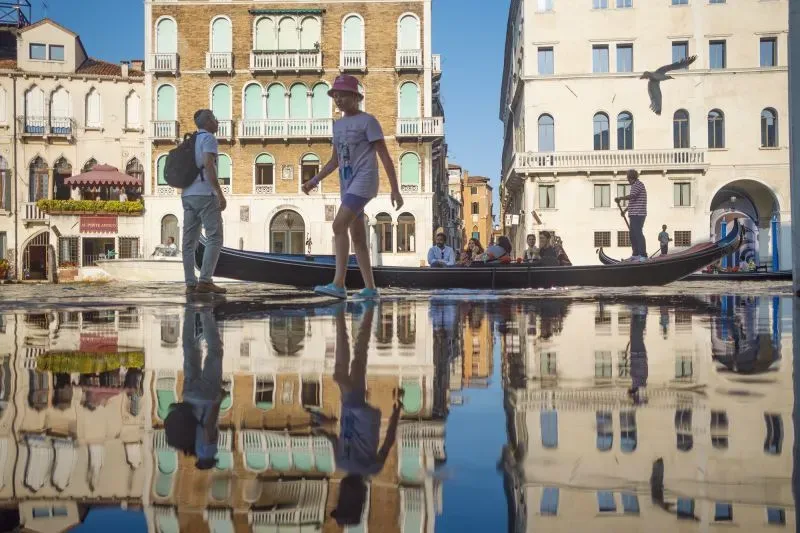 مدينة البندقية تعكس جمالها في المياه، مع أشخاص يمشون بجانب قارب، بينما تعكس المباني التاريخية في البحيرة.