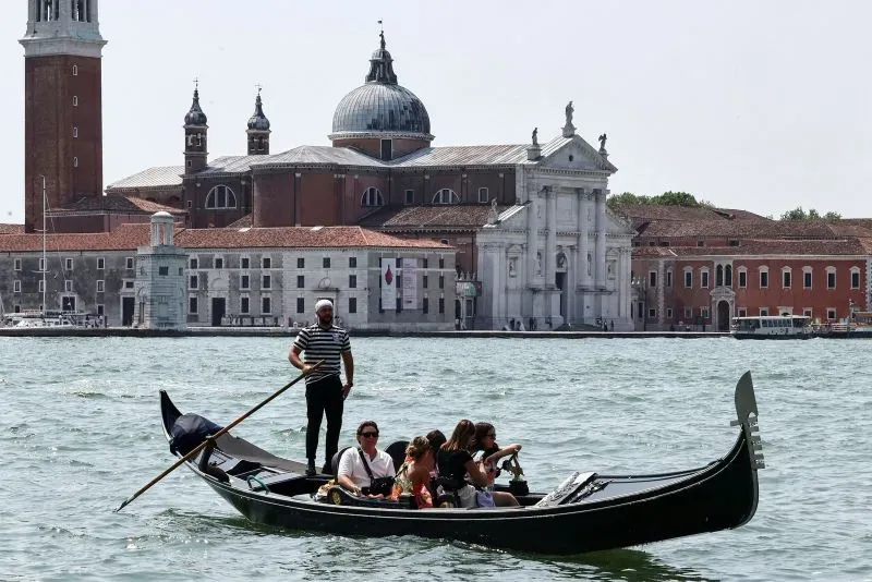 مجموعة من الأشخاص في قارب جندول يتنقلون عبر قنوات البندقية، مع خلفية معمارية تاريخية تبرز جمال المدينة.