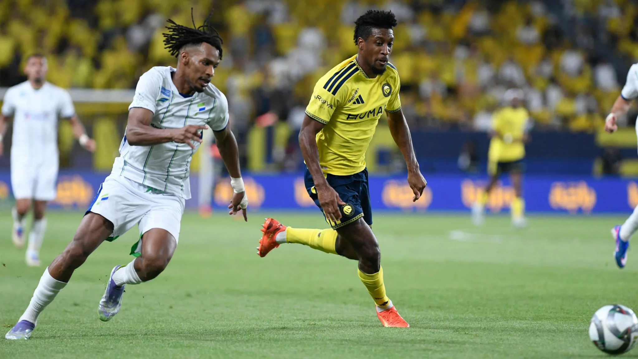 لاعب من النصر يرتدي الأصفر يركض بالكرة متجاوزاً مدافع الفتح في مباراة دوري روشن للمحترفين.