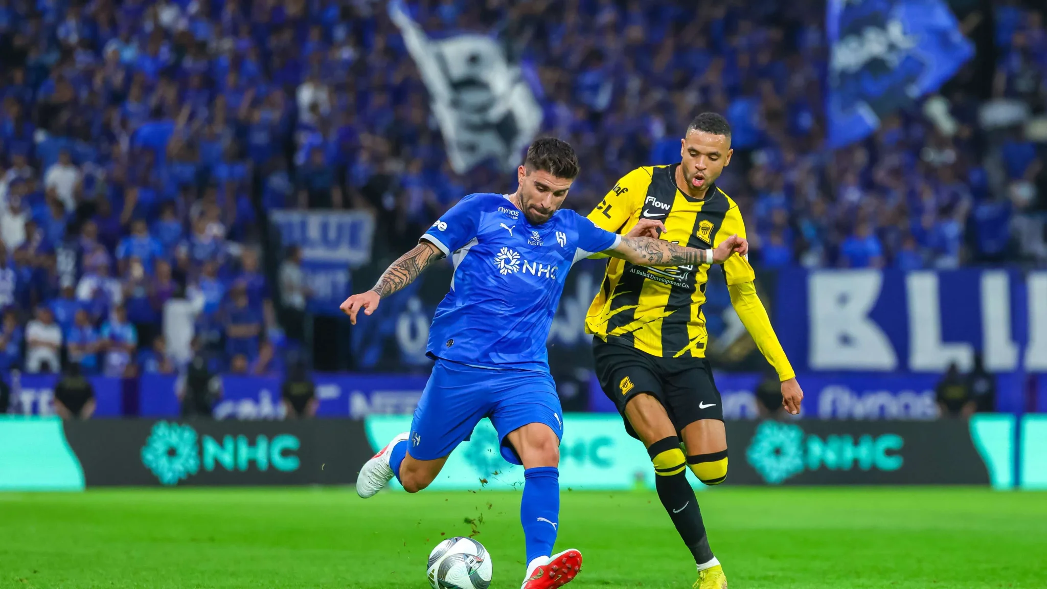لاعبو الهلال والاتحاد يتنافسون على الكرة في مباراة دوري روشن السعودي ضمن كلاسيكو الجولة الثامنة، مع حضور جماهيري كبير.