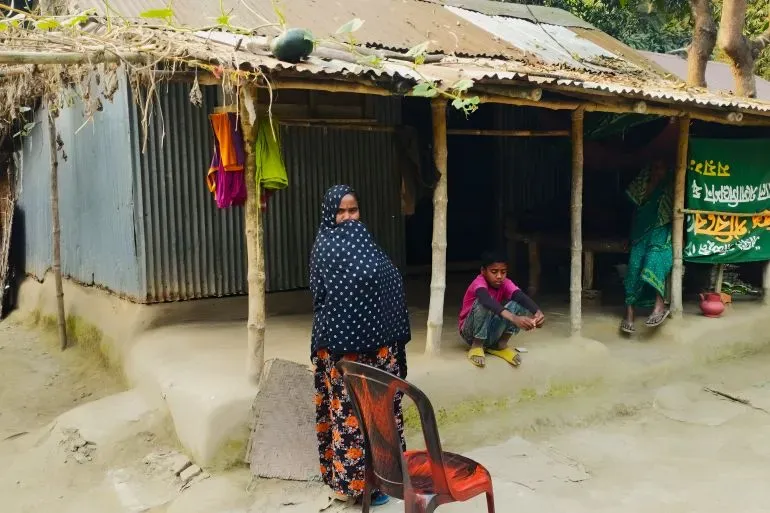 امرأة ترتدي الحجاب تقف أمام منزل بسيط في قرية بايغوني، بينما يجلس طفل بجوارها، مما يعكس التحديات الاجتماعية والاقتصادية في المنطقة.