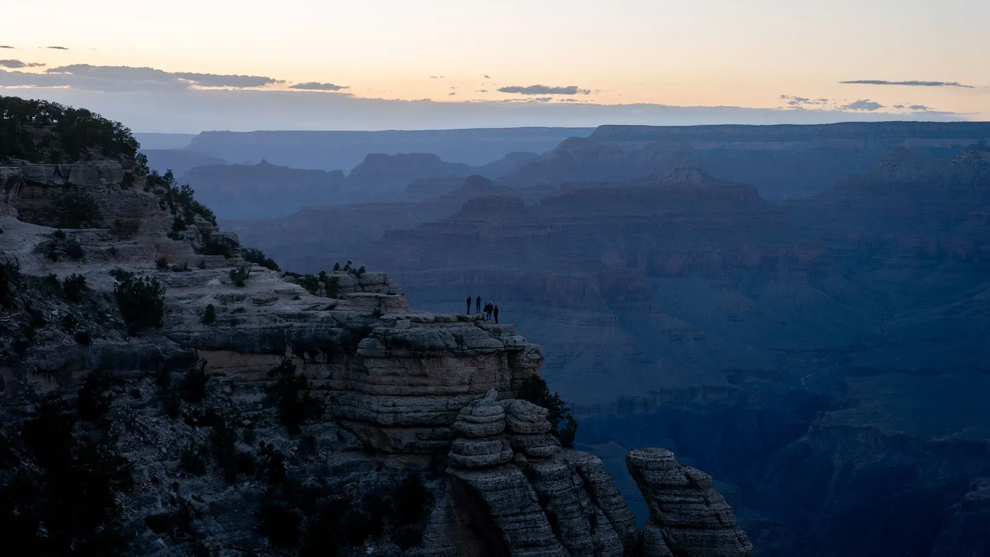 مجموعة من الأشخاص يقفون على حافة صخرية في غراند كانيون خلال غروب الشمس، مع مشهد جبلي رائع في الخلفية.