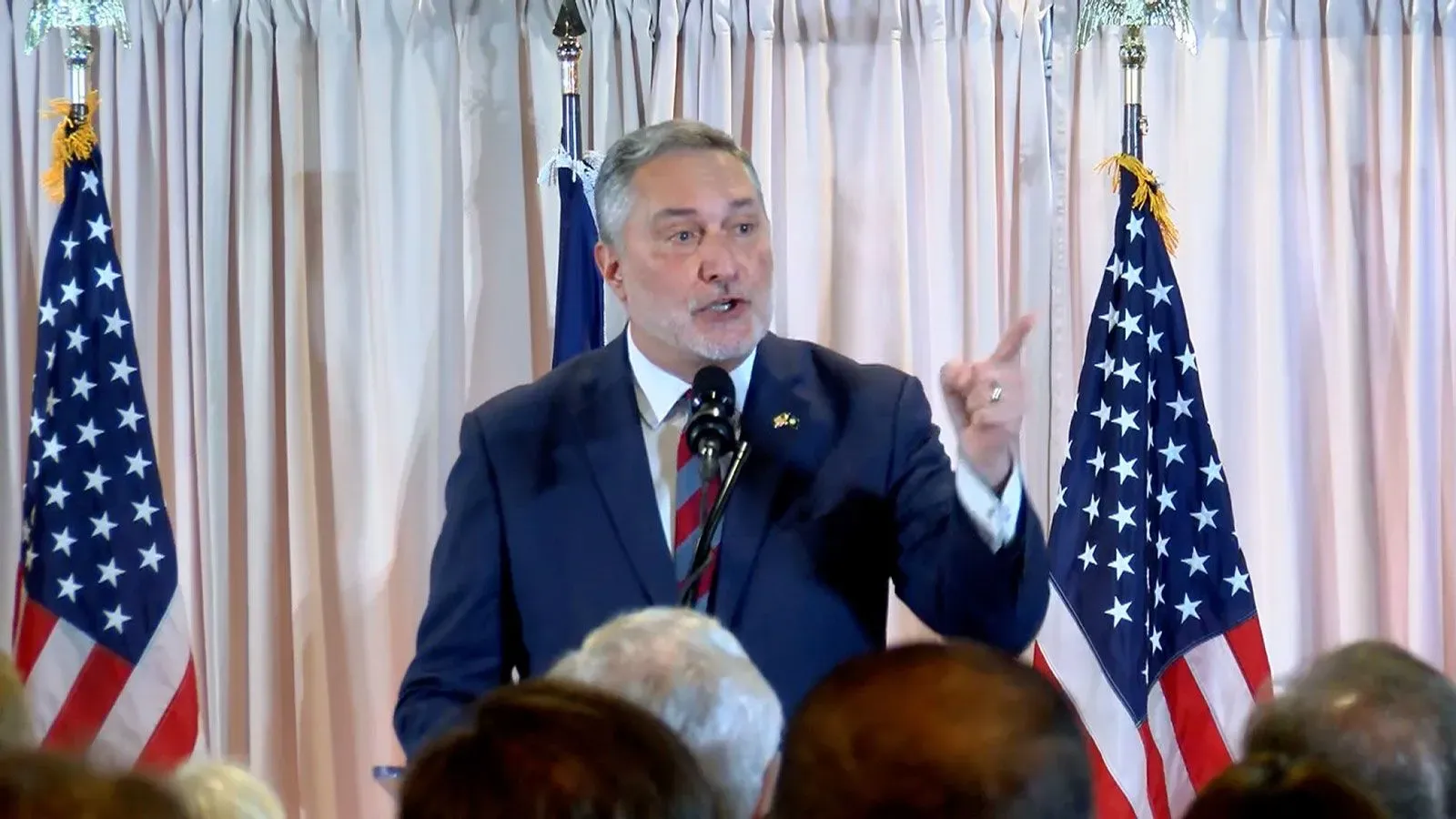 جون ريد، مرشح نائب الحاكم في فيرجينيا، يتحدث أمام جمهور، مع أعلام أمريكية خلفه، في سياق الجدل السياسي حول حساب متنازع عليه.