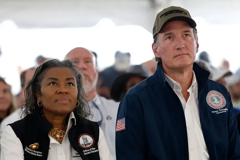 امرأة سوداء ترتدي سترة رسمية، تشاهد حدثًا مع حاكم ولاية فيرجينيا، بينما يبدو الحضور مهتمًا. تعكس الصورة أهمية السباق الانتخابي للمرأة.