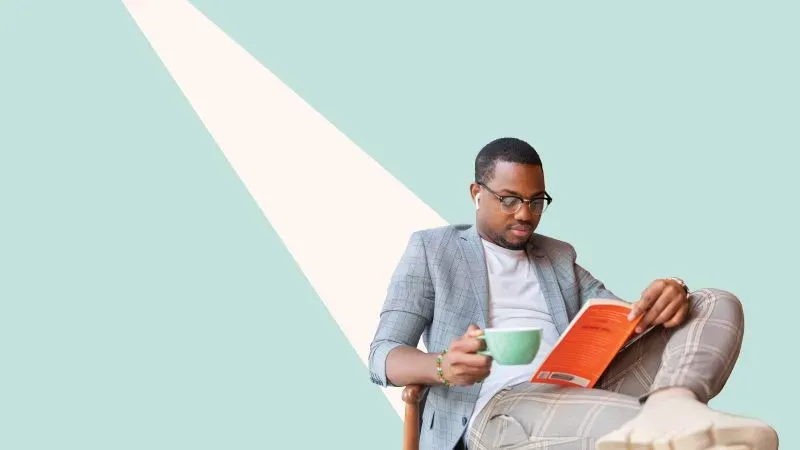 رجل يرتدي سترة رمادية يجلس ممسكًا بفنجان قهوة ويقرأ كتابًا، مضاءً بضوء متسرب من مصدر خارجي.