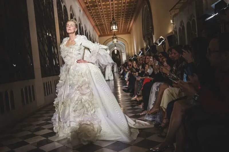 عارضة أزياء ترتدي فستان زفاف مميز بتفاصيل فاخرة وتصميم غير تقليدي، خلال عرض أزياء في برشلونة، مع جمهور يتابع بإعجاب.