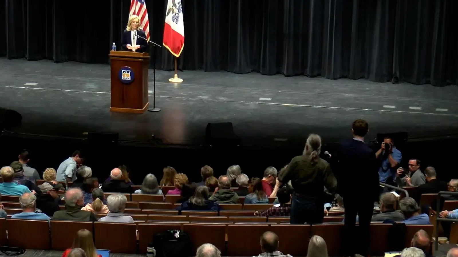 نائبة الكونغرس آشلي هينسون تتحدث في قاعة بلدية في ولاية أيوا، حيث يطرح الحضور أسئلة نقدية حول إدارة ترامب وسط أجواء متوترة.