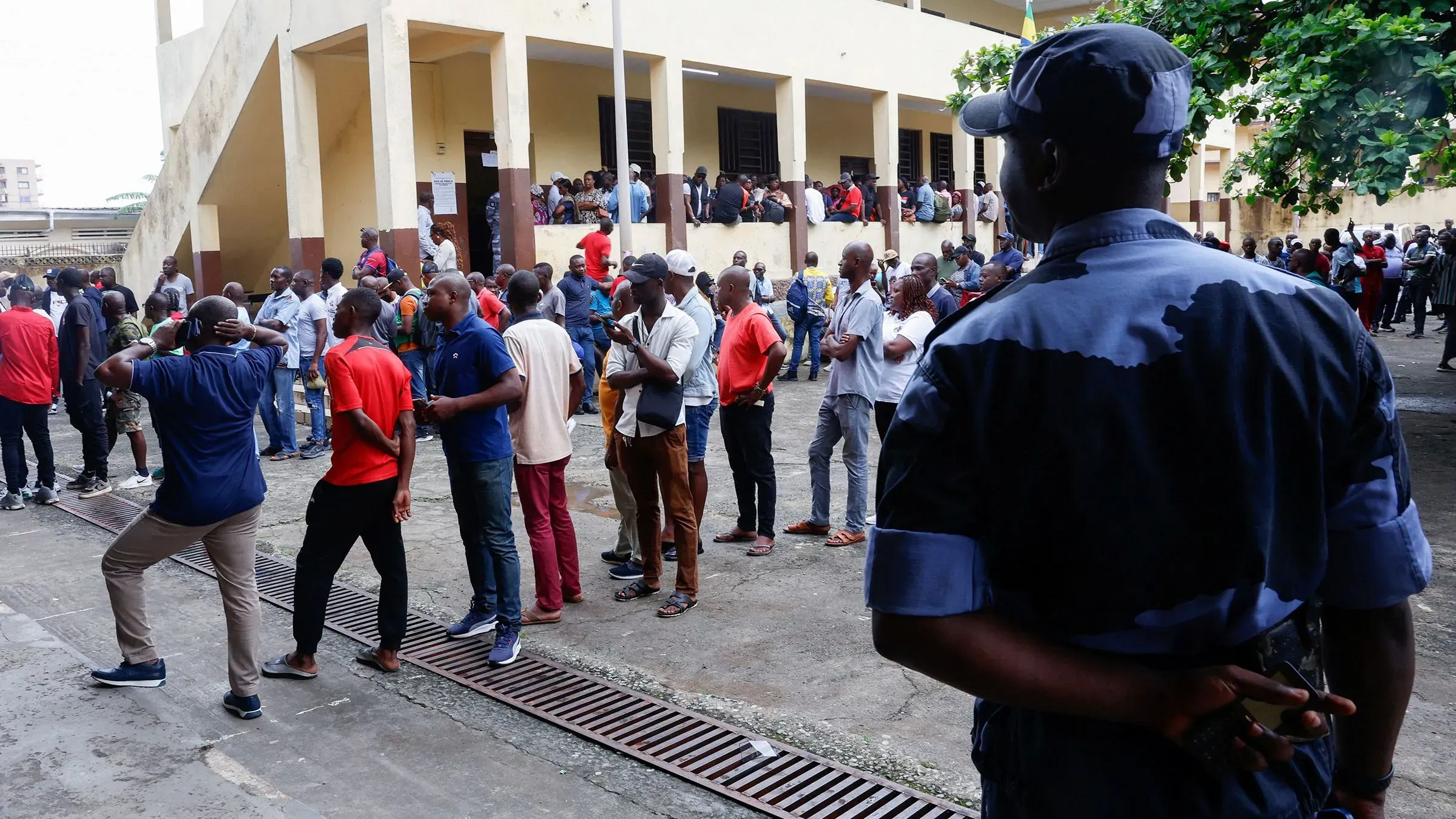 ناخبون من مختلف الأعمار يصطفون في مركز اقتراع بالعاصمة ليبرفيل، بينما يراقبهم ضابط شرطة، في انتخابات رئاسية تاريخية.