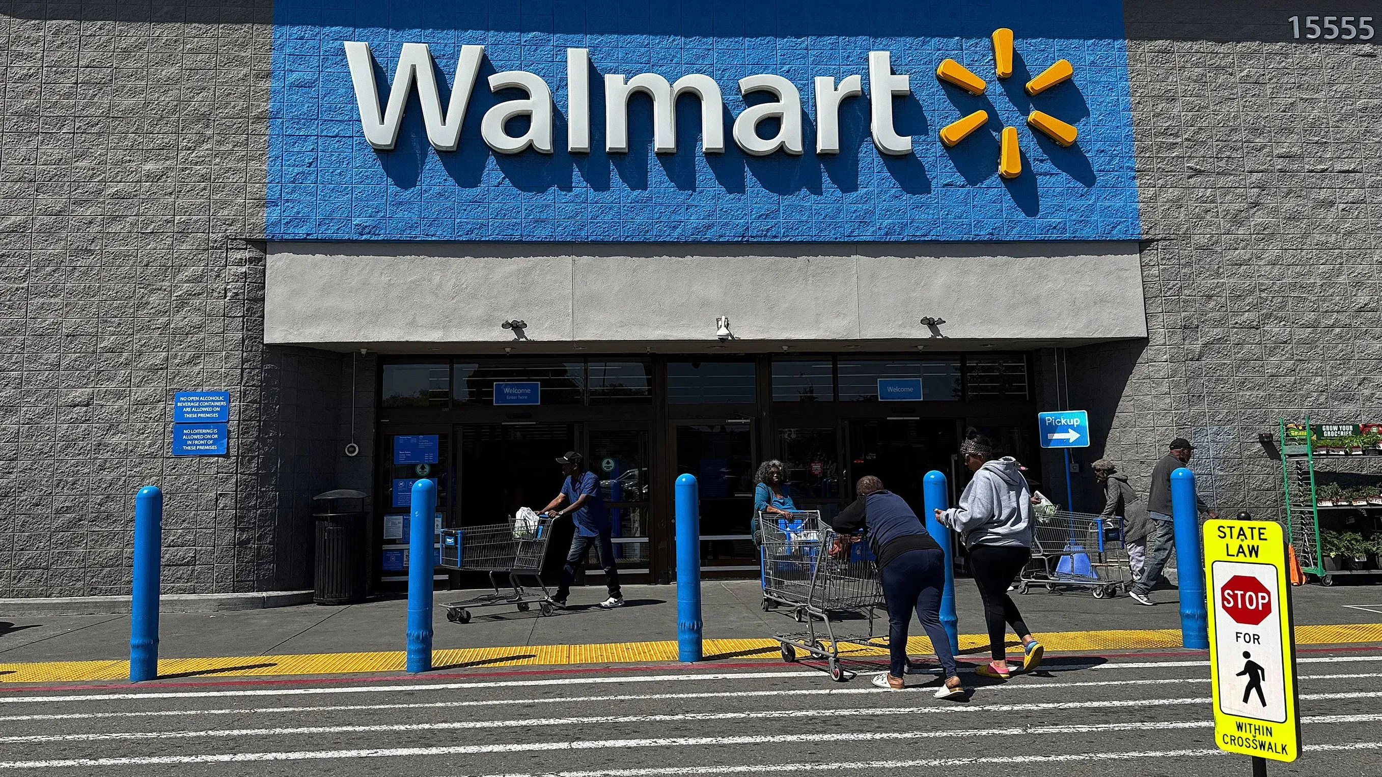 مدخل متجر وول مارت، حيث يتسوق الزبائن مع عربات التسوق، مع وجود لافتة تحذيرية على الرصيف. تعكس الصورة تأثير الرسوم الجمركية على الأسعار.