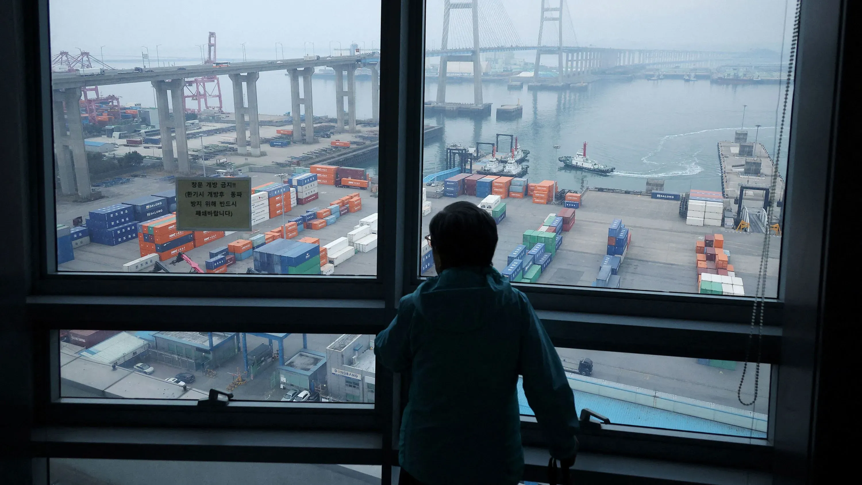 شخص ينظر من نافذة إلى ميناء مزدحم، حيث تتواجد حاويات ملونة وسفن، مما يعكس تأثير التعريفات الجمركية على التجارة.