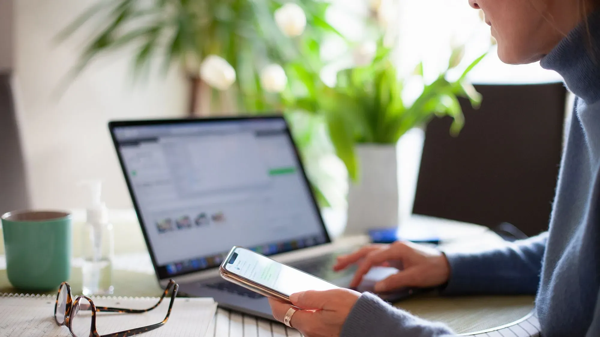 امرأة تستخدم هاتفها الذكي أثناء العمل على الكمبيوتر المحمول في بيئة منزلية مريحة، مع وجود نباتات في الخلفية.