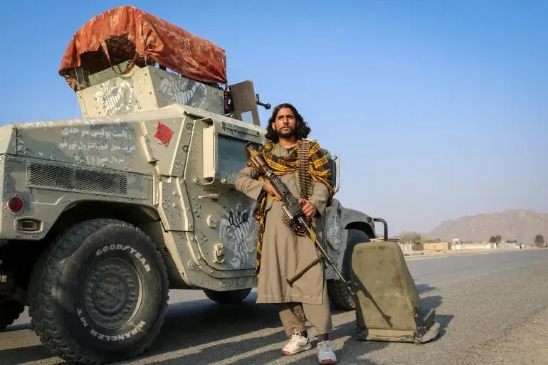مقاتل من طالبان يقف بجانب مركبة عسكرية في منطقة نائية بأفغانستان، مع خلفية جبال وصحراء، يعكس التوترات الأمنية في البلاد.