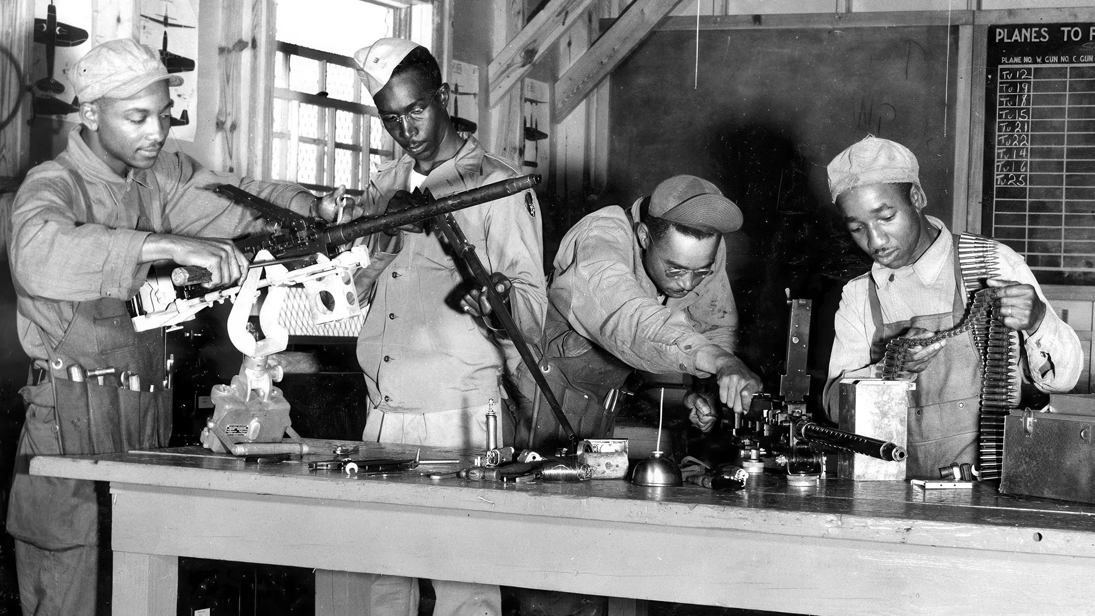 مجموعة من أربعة جنود أمريكيين من أصل أفريقي يعملون على صيانة الأسلحة في ورشة خلال الحرب العالمية الثانية، مع التركيز على مهاراتهم الفنية.