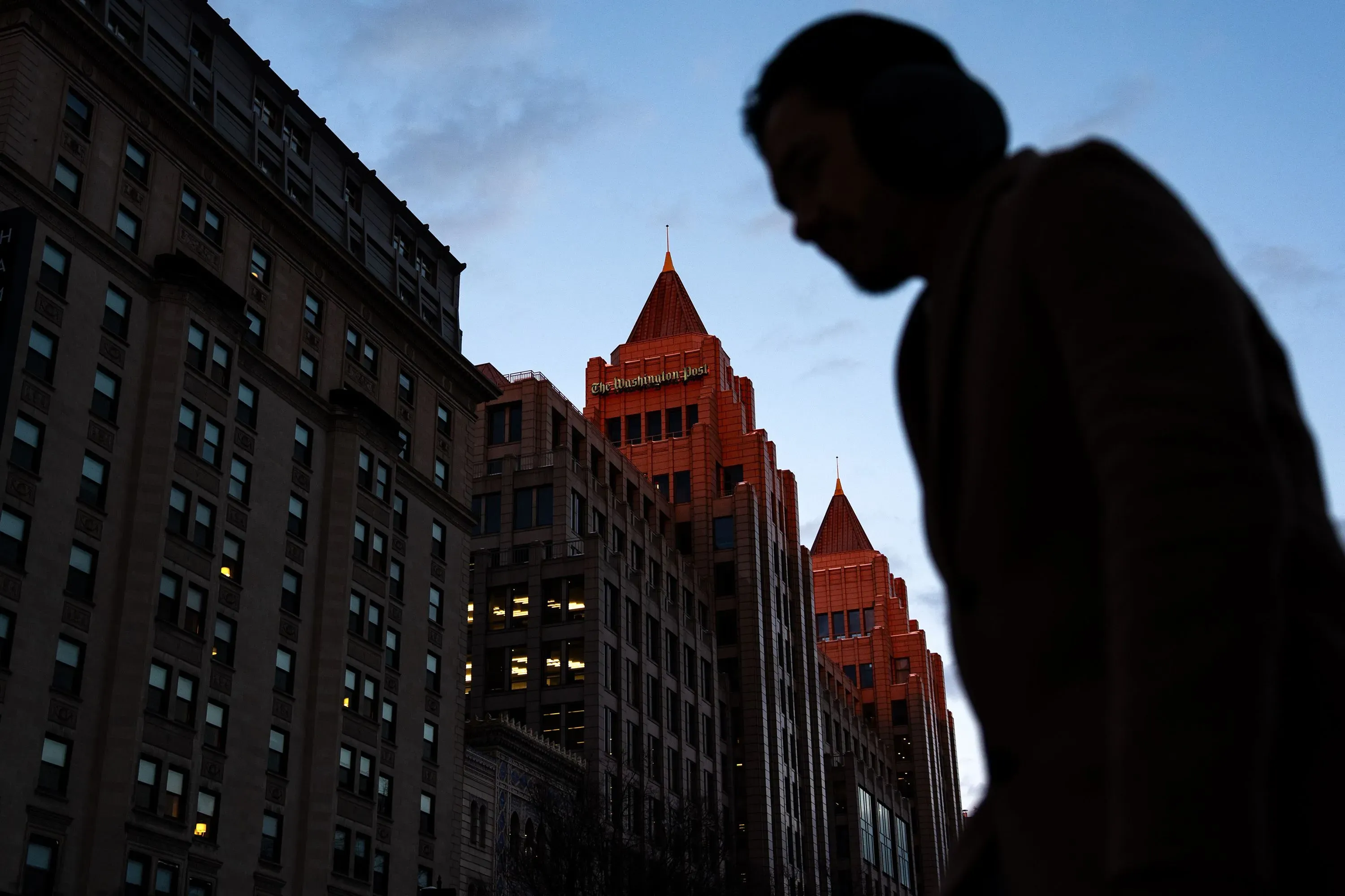رجل يسير بجوار مبنى صحيفة واشنطن بوست، حيث تظهر الأضواء في النوافذ مع غروب الشمس، مما يعكس التوترات الحالية في غرفة الأخبار.
