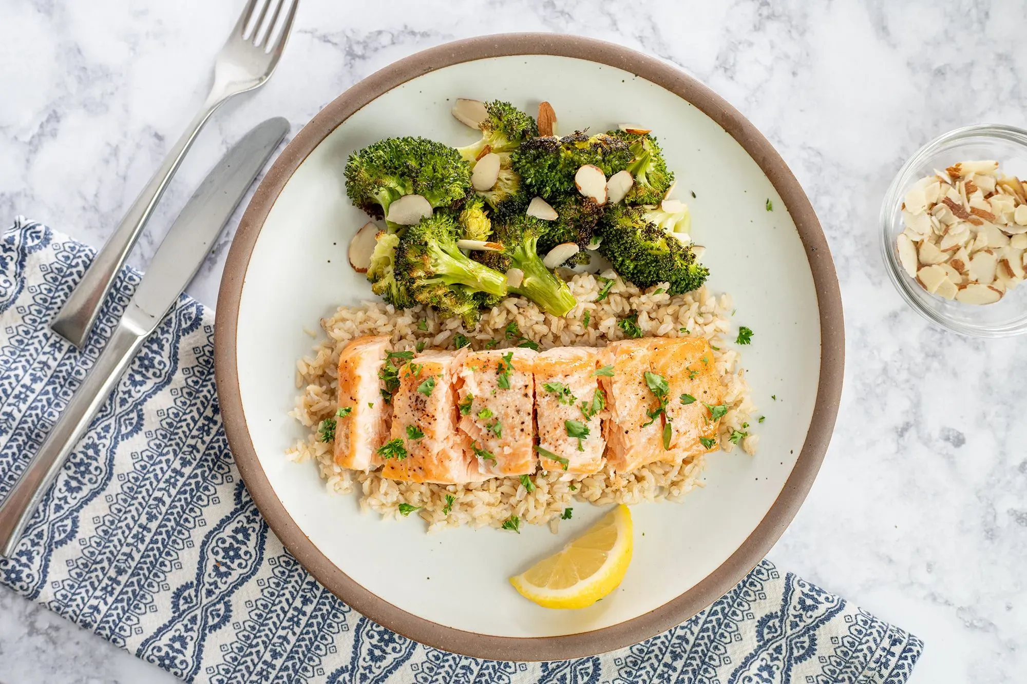 طبق يحتوي على سمك السلمون المشوي مع أرز بني وبروكلي، مزين باللوز وشرائح ليمون، يعكس خيارات غذائية صحية.