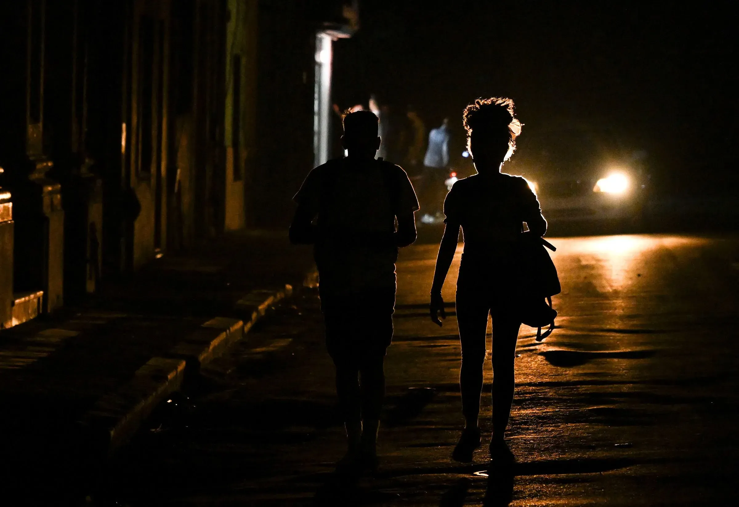 شخصان يسيران في شارع مظلم في هافانا، مع إضاءة خافتة من السيارات، مما يعكس الأجواء الصعبة التي يعيشها الكوبيون.