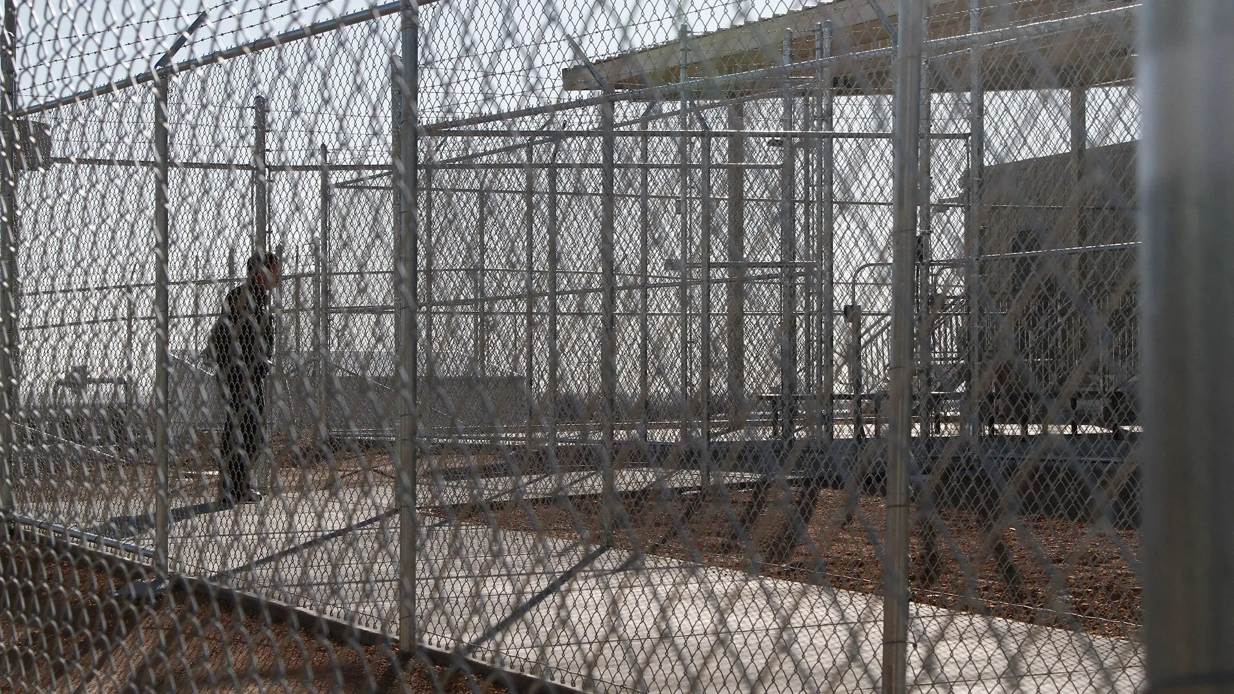 مشهد لمنشأة احتجاز حدودية، حيث يظهر شخص داخل قفص معدني، مما يعكس الظروف القاسية التي يعيشها المهاجرون.