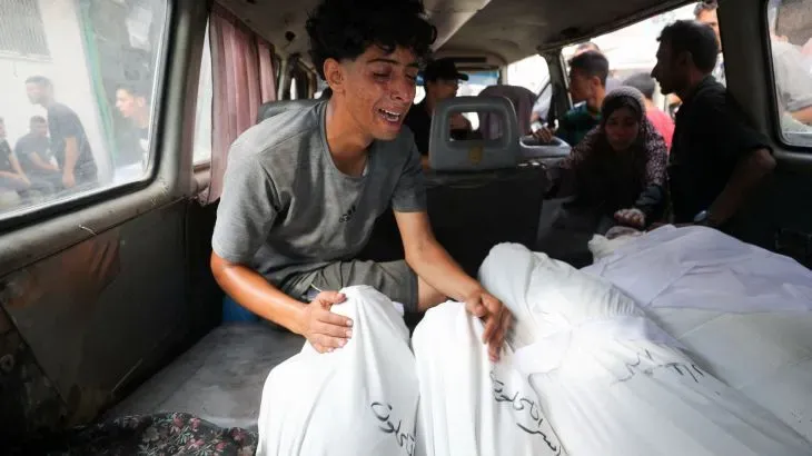 شاب يبكي بجانب جثث مغطاة بأكفان بيضاء داخل سيارة إسعاف في غزة، معبرًا عن الحزن العميق بسبب فقدان أحبائه في الأحداث الجارية.