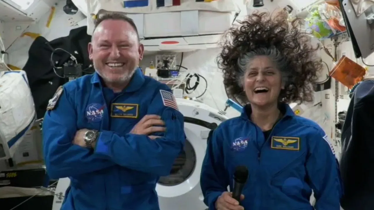 يظهر رائدا الفضاء سوني ويليامز وبوتش ويلمور في محطة الفضاء الدولية، يبتسمان ويتحدثان، مع أدوات فضائية خلفهما.