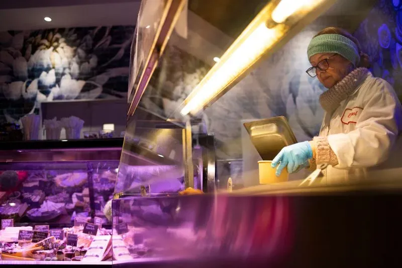 امرأة ترتدي سترة بيضاء وقبعة صوفية، تقوم بتحضير الطعام في متجر، مع خلفية مزينة بالزهور.