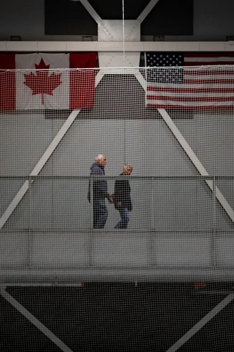 زوجان يمشيان على جسر في مركز رياضي، مع وجود علمي كندا والولايات المتحدة في الخلفية، يعكسان التوترات التجارية بين البلدين.