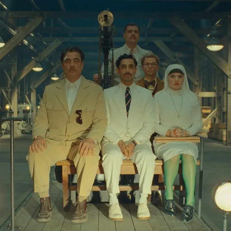 مجموعة من الشخصيات الرئيسية من فيلم "المخطط الفينيقي" لويس أندرسون، يجلسون في صف، يرتدون ملابس أنيقة تعكس أسلوب الفيلم الفريد.