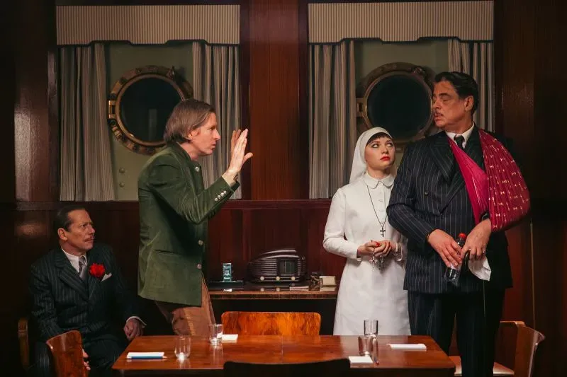 مشهد من فيلم "المخطط الفينيقي" لويس أندرسون، يظهر شخصيات تتفاعل في غرفة ذات طابع كلاسيكي، مع تفاصيل ملابس أنيقة.