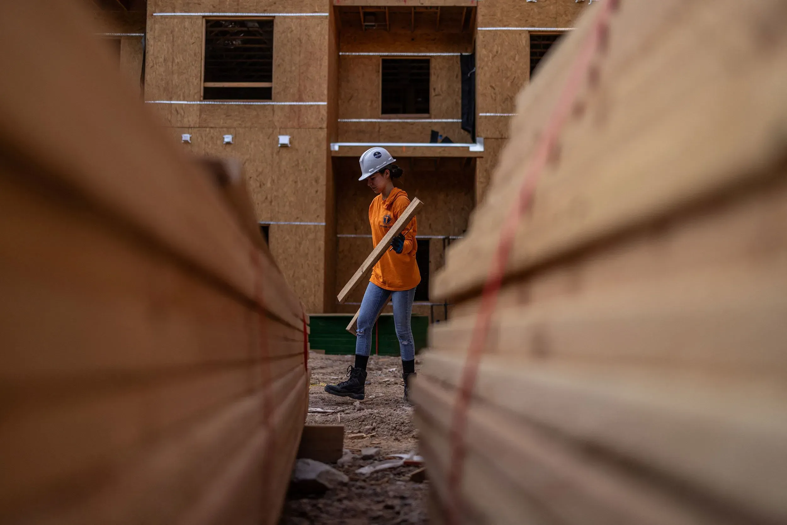 شابة تحمل قطعة خشب في موقع بناء، مع وجود صفوف من الخشب في المقدمة وخلفية تظهر المبنى قيد الإنشاء.