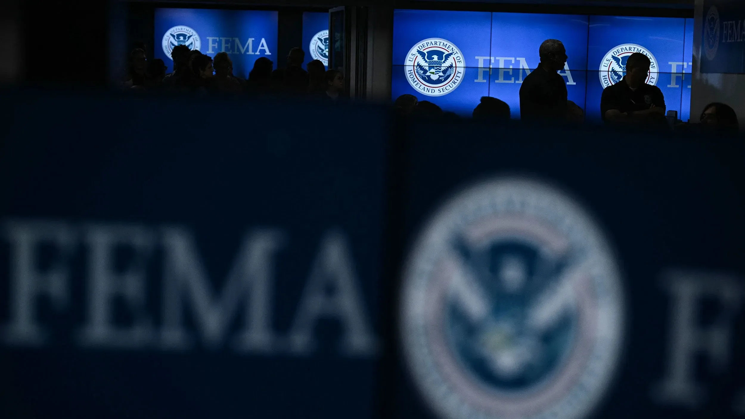 تظهر صورة لموظفين في وكالة إدارة الطوارئ الفيدرالية (FEMA) أثناء اجتماع، مع خلفية تحمل شعار الوكالة. تعكس الصورة حالة عدم اليقين في الاستجابة للكوارث.