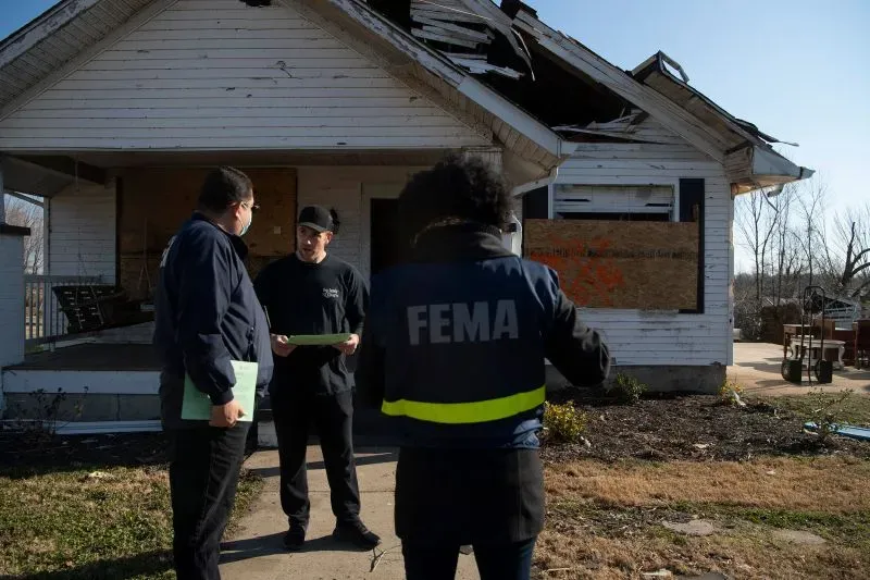 موظفان يتحدثان أمام منزل متضرر من الكوارث، بينما يرتدي أحدهما سترة تحمل شعار إدارة الطوارئ الفيدرالية (FEMA)، مما يعكس التحديات في الاستجابة للكوارث.