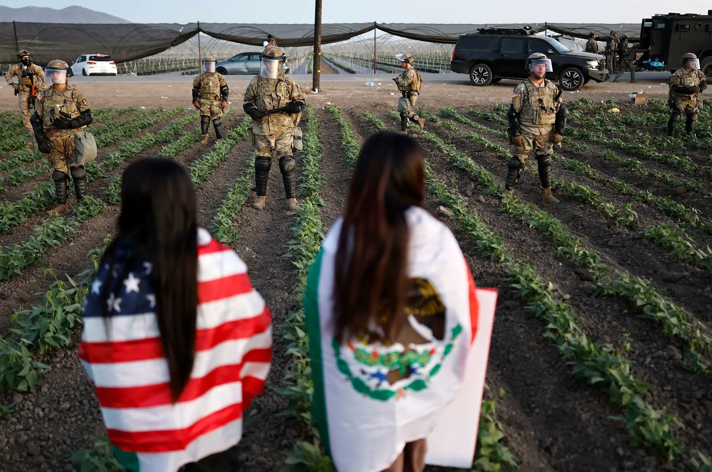 جنود يرتدون زيًا عسكريًا يراقبون حقلاً زراعيًا، بينما تقف امرأتان تحملان علمي الولايات المتحدة والمكسيك.