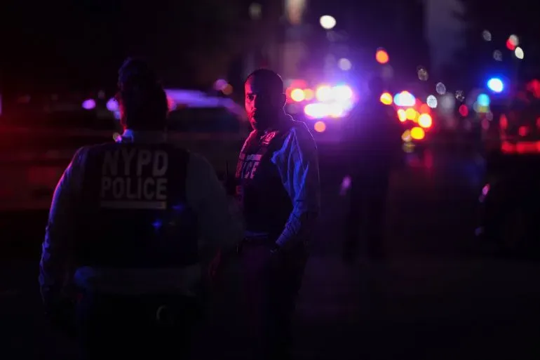 رجال شرطة من إدارة شرطة نيويورك في موقع حادث إطلاق نار، مع أضواء سيارات الطوارئ اللامعة في الخلفية، بعد وقوع حادث مأساوي في مانهاتن.