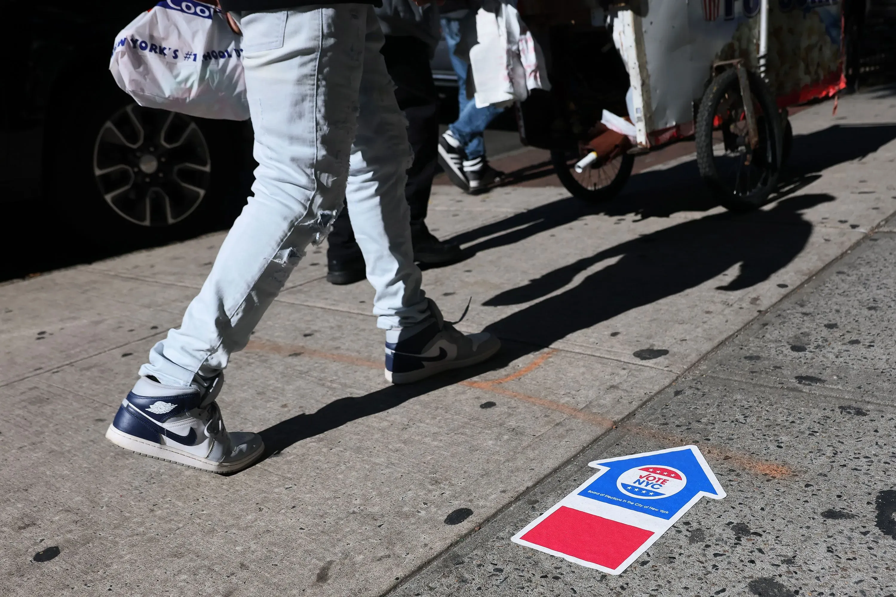 أشخاص يسيرون على الرصيف بالقرب من علامة تشير إلى التصويت، تعكس أهمية المشاركة في الانتخابات المقبلة في نيويورك.