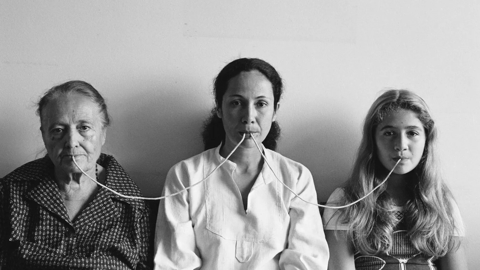 صورة بالأبيض والأسود لثلاث نساء من أجيال مختلفة يجلسن معًا، ويمتد خيط بين أفواههن، مما يعكس موضوع التواصل بين الأجيال.