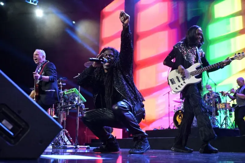 عازف الجيتار ماركوس ميلر يؤدي عرضًا حيًا مع فرقة موسيقية، بينما تضيء خلفهم أضواء ملونة، مع تركيز على حيوية موسيقى الفانك.