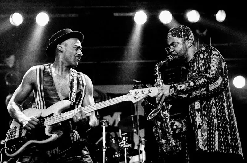 عازف الجيتار ماركوس ميلر يعزف في عرض حي، بينما يعزف عازف الساكسفون بجانبه، وسط أضواء المسرح.