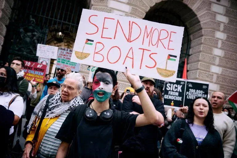 متظاهر يحمل لافتة مكتوب عليها "أرسلوا المزيد من القوارب" خلال احتجاج لدعم غزة، بينما يظهر على وجهه ألوان العلم الفلسطيني.