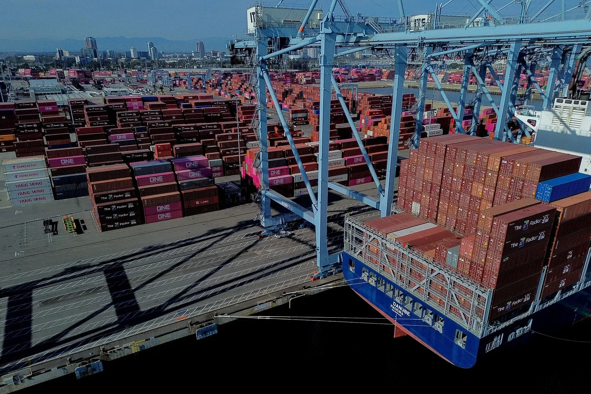 صورة لميناء مزدحم يظهر فيه حاويات شحن ملونة مكدسة على أرصفة الميناء، مع سفينة شحن تحمل حاويات جديدة.