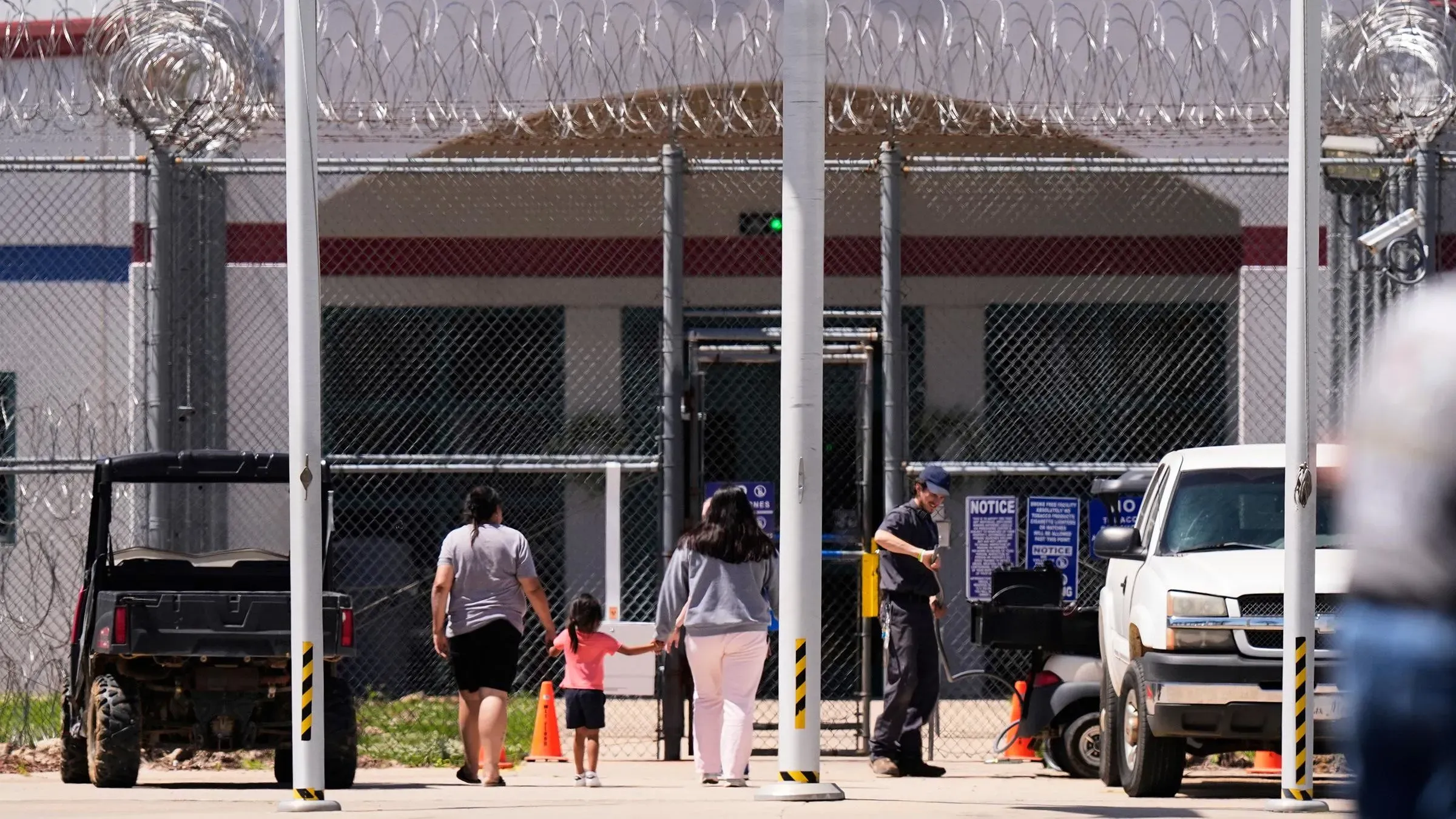 امرأتان تحملان أطفالًا صغيرين، تسيران نحو مركز احتجاز محاط بسياج شائك، مما يعكس قضايا الترحيل العائلي في الولايات المتحدة.